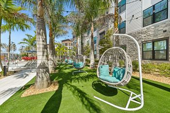 A white swing chair with a green cushion is in the foreground of a sunny outdoor area with palm trees and a building in the background.